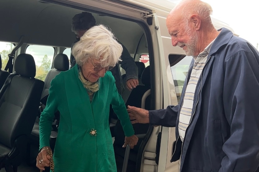 An older balding man helps an elderly woman dressed in green and scarf around her neck off a bus.