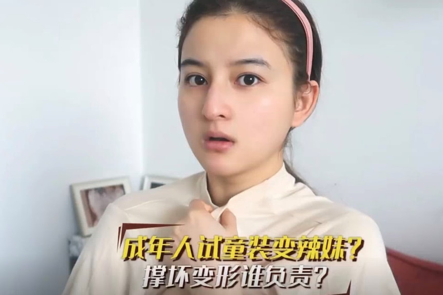 A young woman closes the collar of her top near her neck as she looks into a camera and speaks.