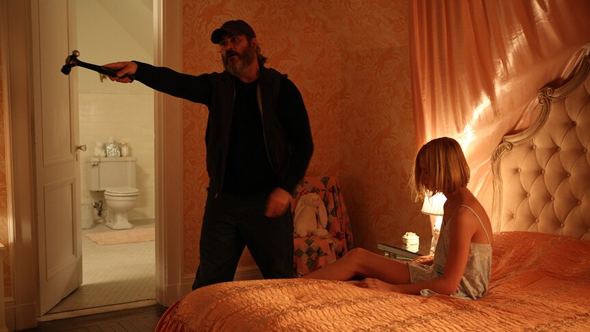 Joaquin Phoenix standing and pointing with hammer while Ekaterina Samsonov sits on bed in film You Were Never Really Here.