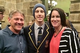 A student in a school blazer and beanie flanked by his parents.