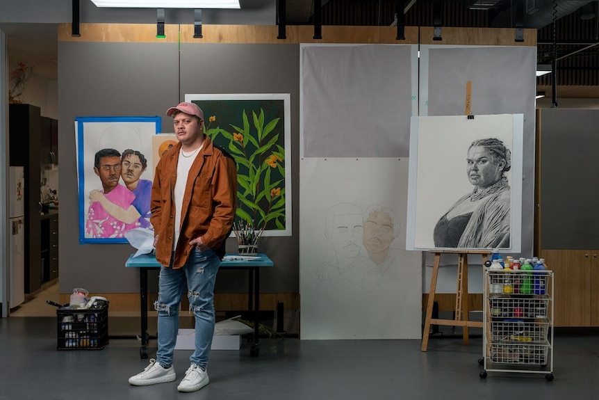 Young Indigenous man wears denim jeans, a pink cap and an ochre jacket in an art studio.