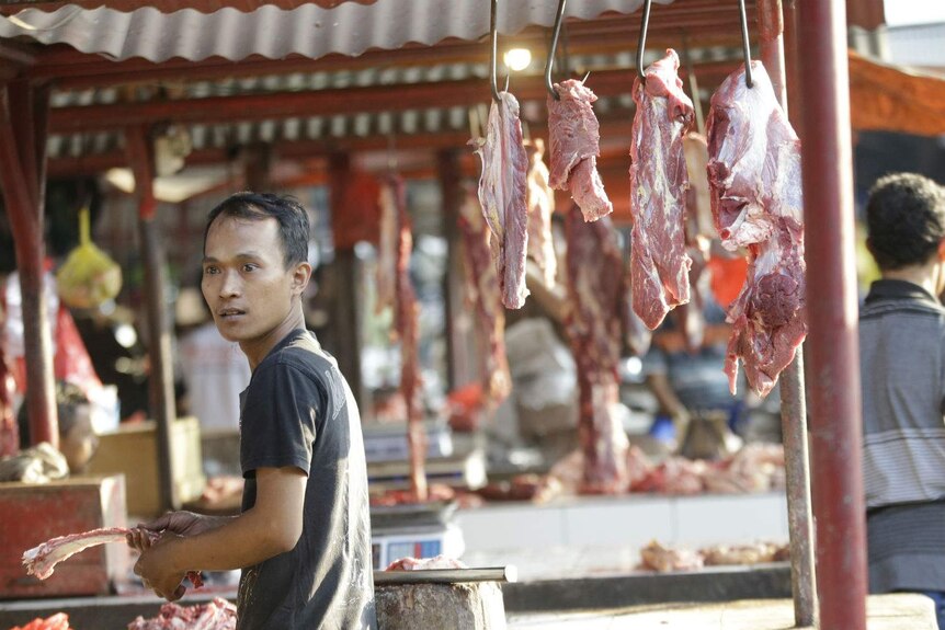 A man holds an animal bone at an open butcher's stall at an indoor/outdoor wet market.