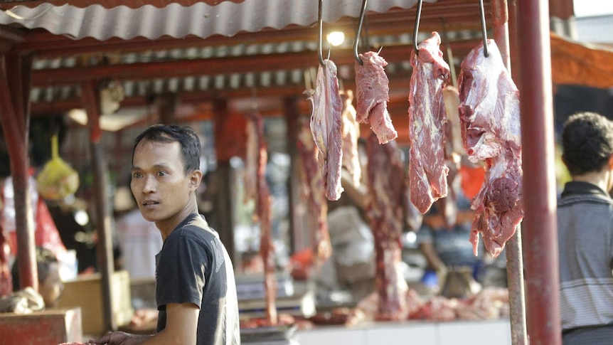 A man holds an animal bone at an open butcher's stall at an indoor/outdoor wet market.