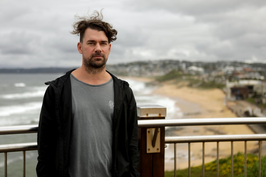 A windswept man in a hoodie poses on a bluff above a beach