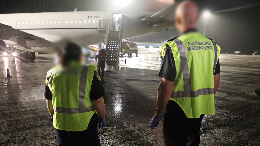 Two Australian Border Force officers in high-vis vests stand near a plane on the tarmac at night.