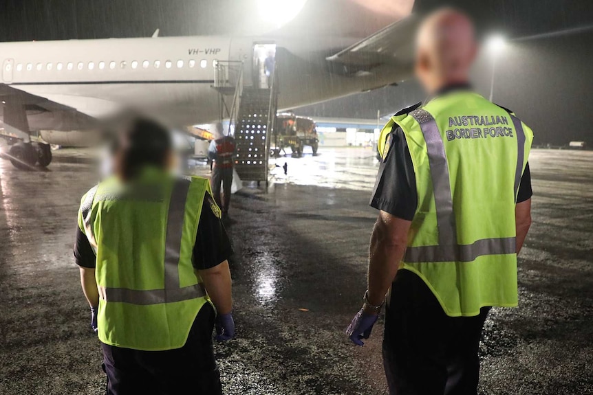 Two Australian Border Force officers in high-vis vests stand near a plane on the tarmac at night.