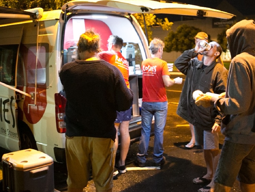 Four homeless men and two volunteers standing behind van in various poses