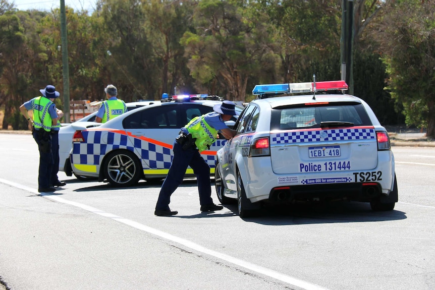 A police officer leans his elbows on the window of a police car on the road, in front of more police cars and officers.