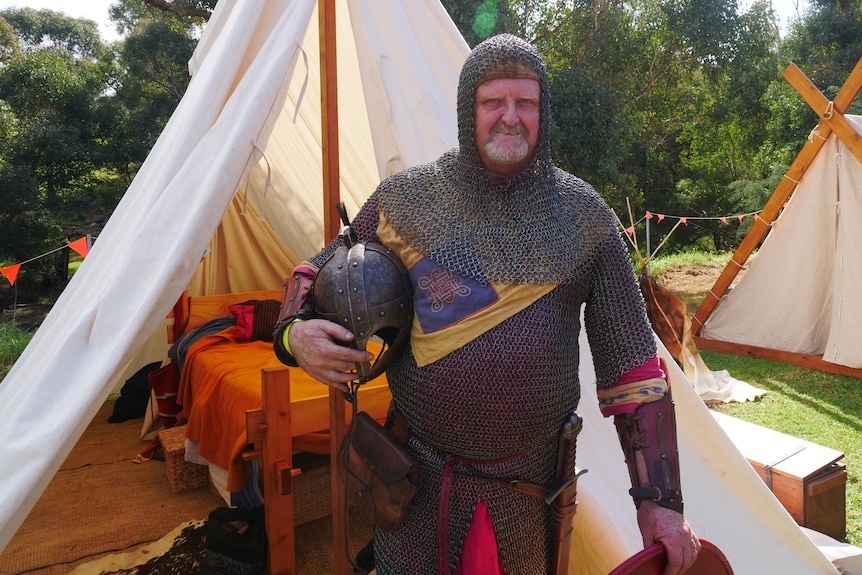 An older man stands in front of a cloth tent wearing a suit of armour.