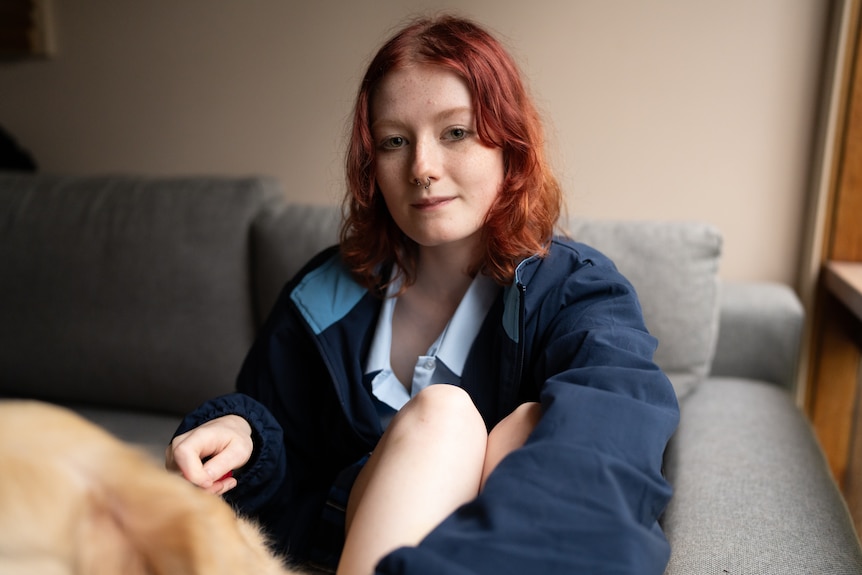 A teenage girl sits on a couch, looking at the camera, with a serious, reflective expression.