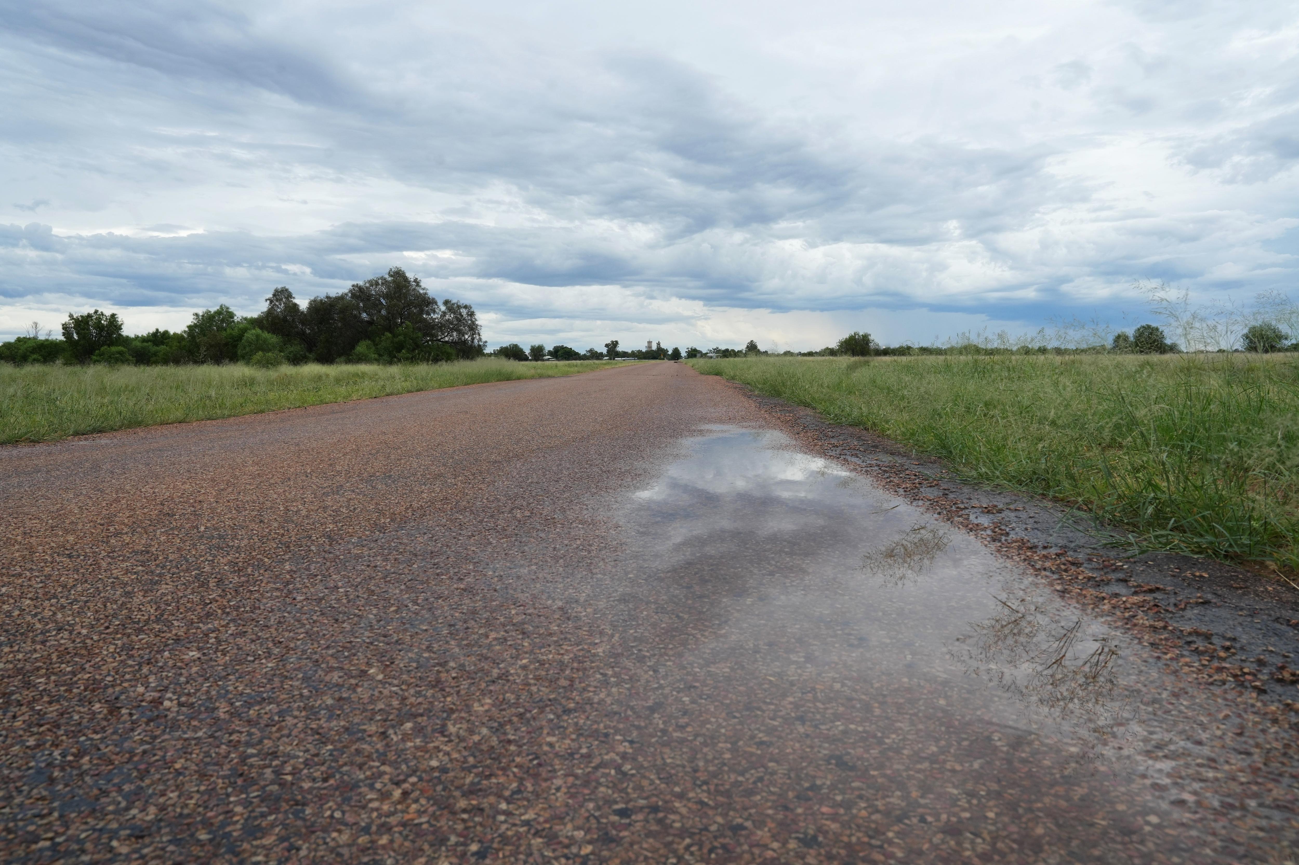 Un charco de agua en un camino rural que refleja la hierba y un cielo gris. 