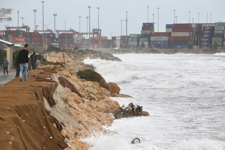 Man looks over embankment at churning sea crashing against rocks. Shipping containers in background.