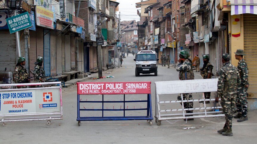 You look down a long street in Indian-controlled Kashmir, with a military roadblock in the foreground flanked by officers.