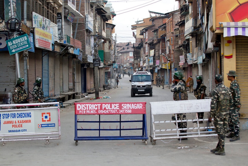 You look down a long street in Indian-controlled Kashmir, with a military roadblock in the foreground flanked by officers.