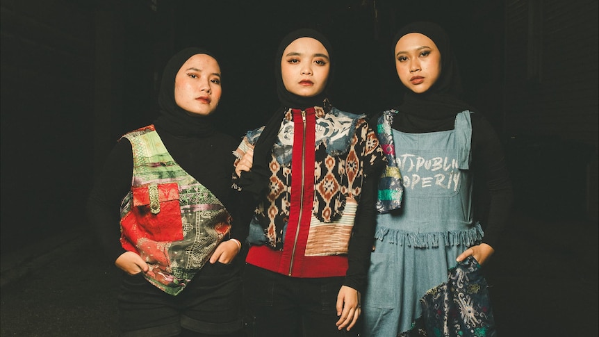 three women in colourful urban wear and black hijabs stand confidently against a black background.