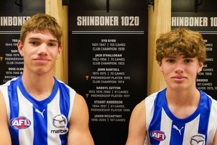 Two teenage boys in football guernseys sit smiling on a changeroom bench. Player names including are on the wall behind them.