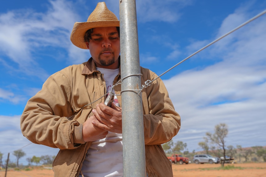 A stockman wearing a straw Akubra fixes a fence.