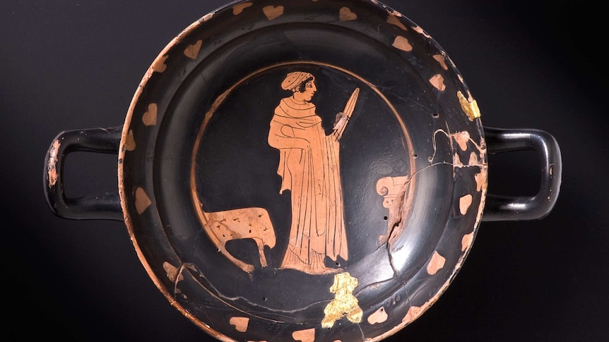 View from above of a Greek drinking vessel. It is black with two small handles. Orange figure with sword in the centre of design