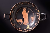 View from above of a Greek drinking vessel. It is black with two small handles. Orange figure with sword in the centre of design