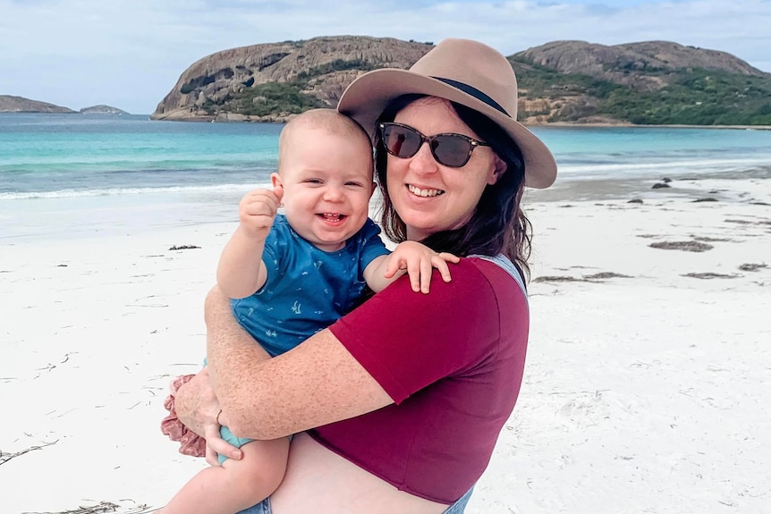 Woman stands on a beautiful beach, holding her laughing baby boy.