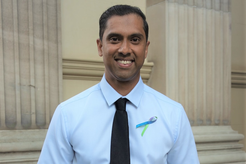 Man in white shirt and dark tie standing with hands in pockets smiling.