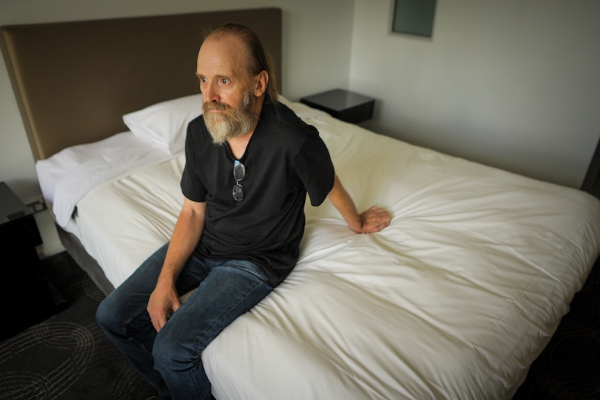 Older man wearing a black top and jeans, sitting on a bed.