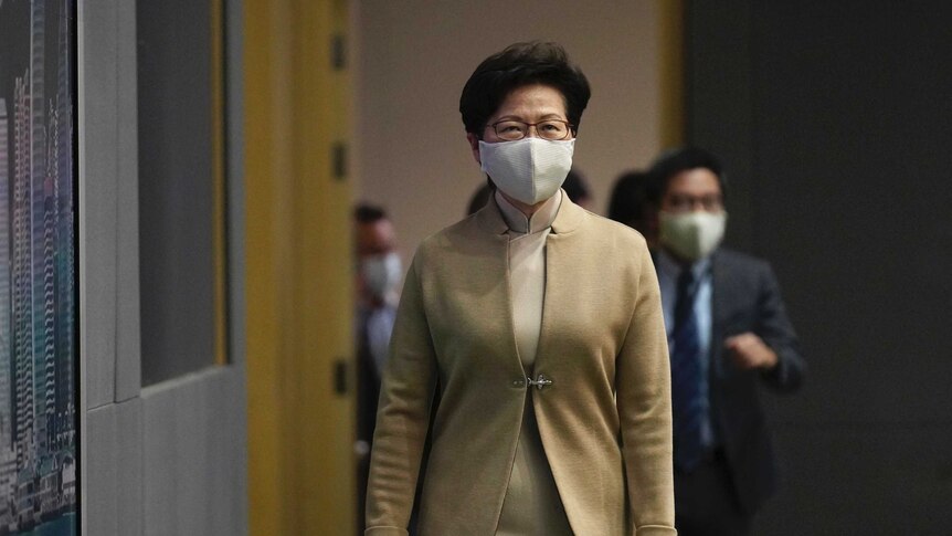 A woman with dark hair and wearing glasses and a face mask walks carrying a blue folder