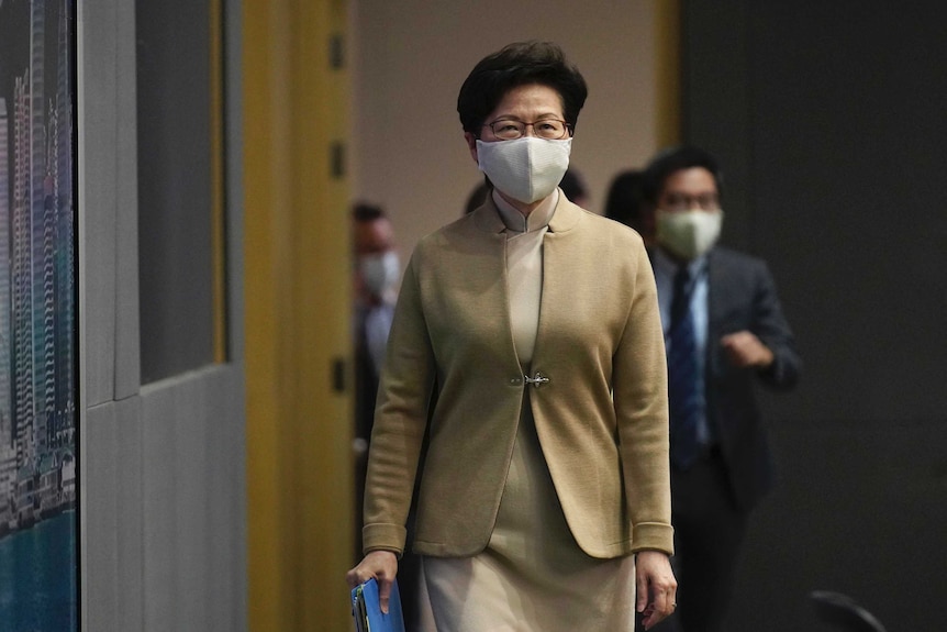 A woman with dark hair and wearing glasses and a face mask walks carrying a blue folder