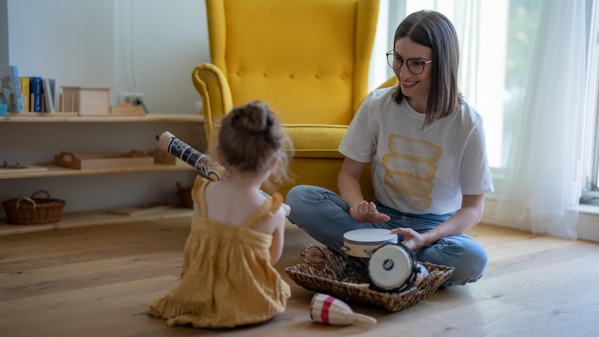 Near a bright yellow armchair, a smiling woman and child sit on the floor near various instruments, like tambourines.