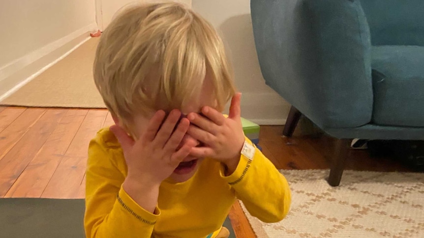 A young child in yellow crouches down, covering his face with his hands.