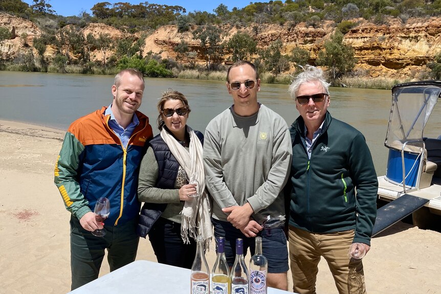 Cuatro personas sonríen, sosteniendo copas de vino a orillas del río Murray, junto a un barco frente a acantilados de piedra caliza.