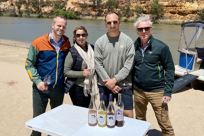 Four people stand smiling, holding wine glasses on the banks of the River Murray by a boat in front of limestone cliffs.