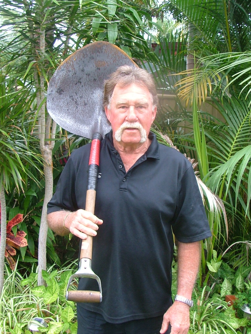 A man with a large moustache and wearing a black t-shirt stands holding a large scoop-shaped shovel against his shoulder.
