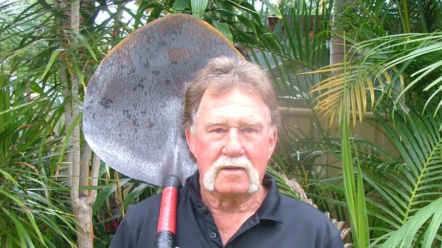 A man with a large moustache and wearing a black t-shirt stands holding a large scoop-shaped shovel against his shoulder.