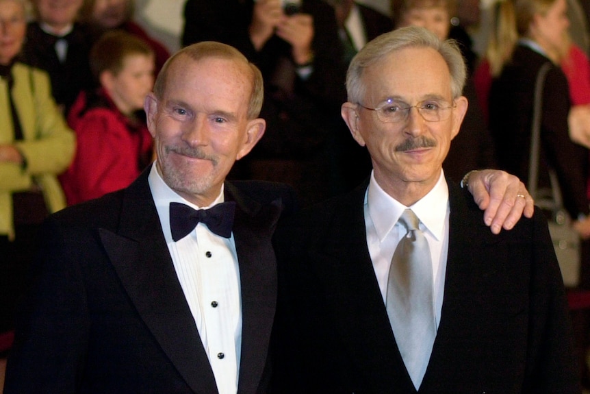 Two men stand with an arm over each other's shoulder. They are both wearing suits.