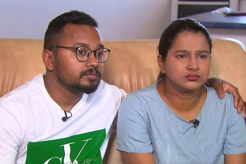 A man in a white shirt with glasses and a woman with a blue shirt sit on a leather couch.