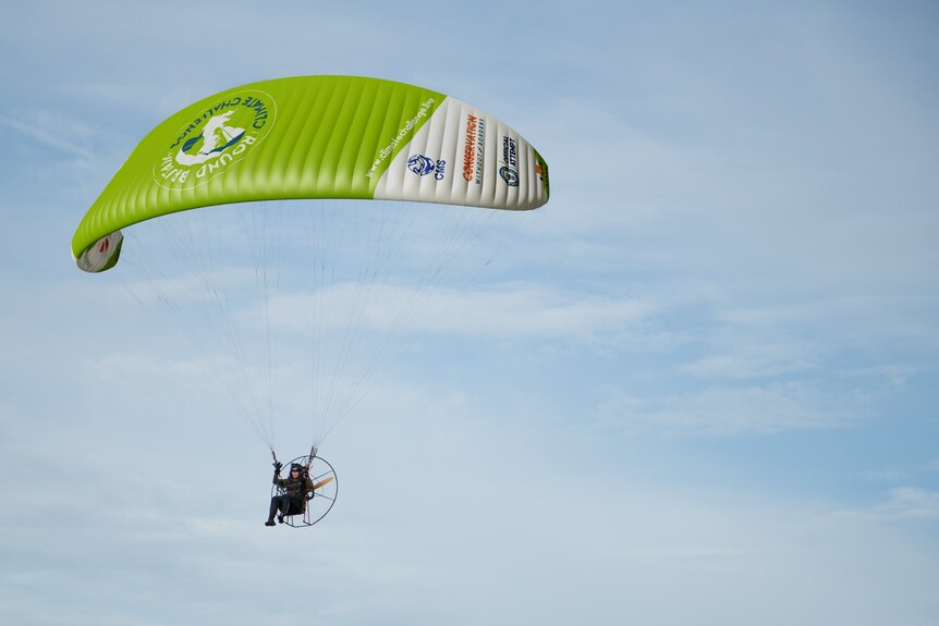 A woman in the air in a paramotor, with a large green and white parachute.