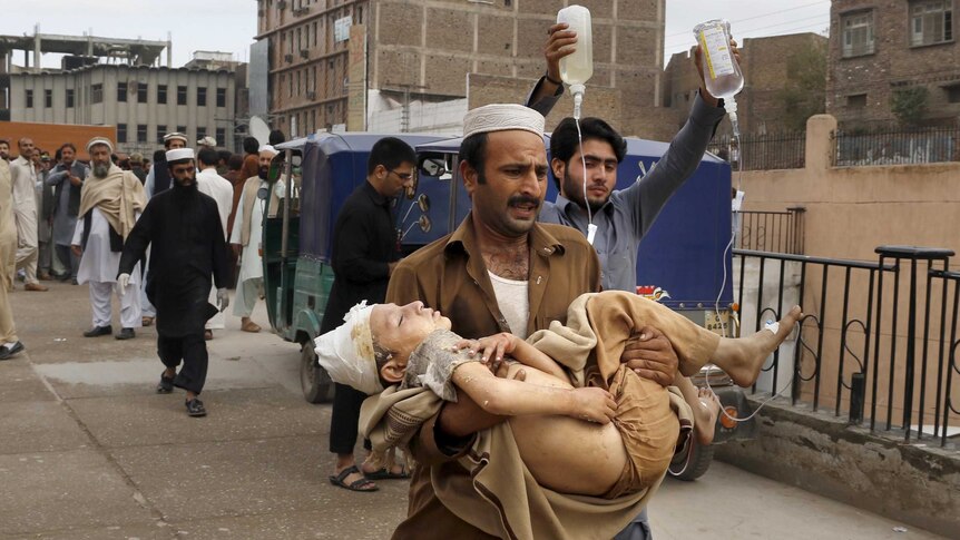 A man carries a Pakistani boy injured in the earthquake as another man walks alongside holding two drips.