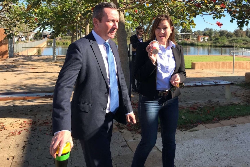 Premier Mark McGowan and Tania Lawrence hold reusable coffee cups while walking under a poinciana tree.