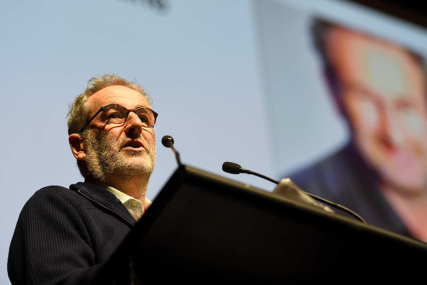 Jon Faine speaks at a lecturn, with a picture of his face projected on the screen behind him.