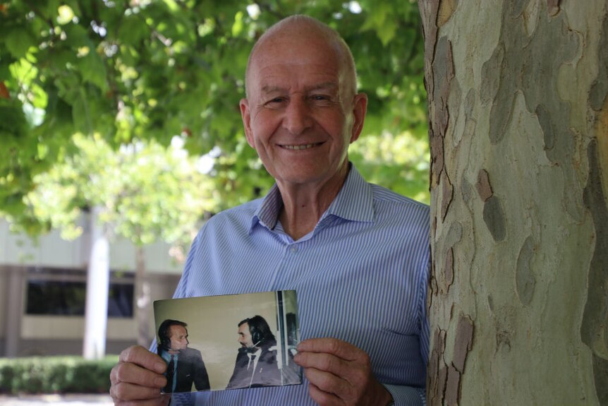 John O'Connell leaning on the trunk of a leafy tree holding an old photograph.
