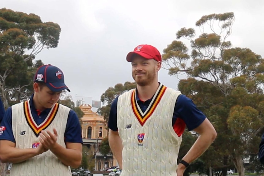 Man stands in cricket whites wearing cap as another man applauds.