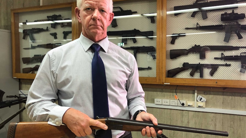 Peter McErlain has a stern look on his face as he holds a rifle and stands in front of a cabinet of guns.