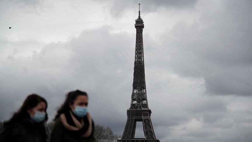 Two women wearing masks walk in front of the Eiffel tower