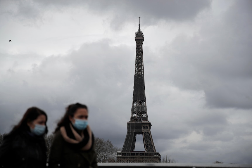 Two women wearing masks walk in front of the Eiffel tower