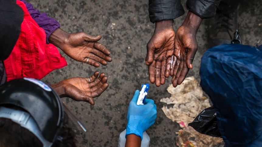 Two pairs of hands are held out with a person spraying hand sanitiser. Photo shot from above looking down.