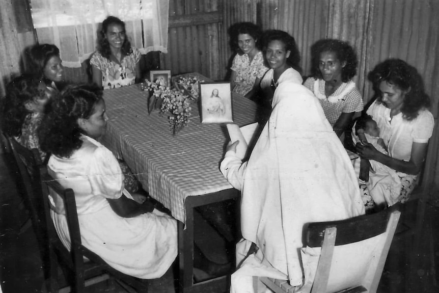 A group of young Indigenous women sit around a table with a nun who is holding a picture of Jesus up