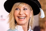 Olivia Newton-John smiles, wearing a mortar board hat
