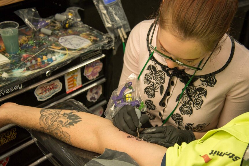 Tattooist Ash Chain presses the needle of her machine against the skin of a man's inner bicep.