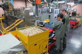 Two women in olive green uniforms and safety goggles stand next to a large crate of golden bullet shells in a factory setting.